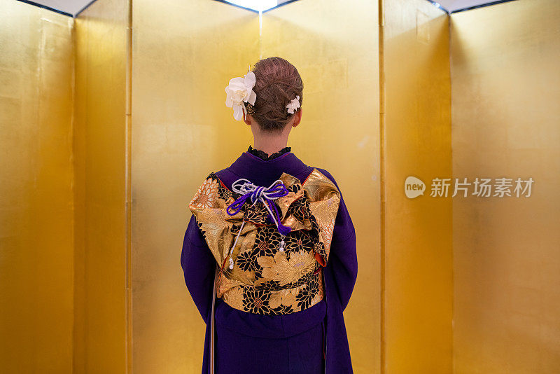 身着“Furisode”和服的日本妇女站在“Seijin Shiki”成人礼的金色“Byo-bu”折叠屏风前的后视图